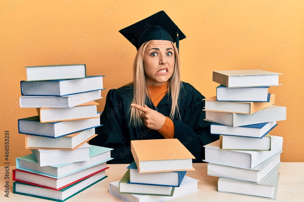 Young caucasian woman wearing graduation ceremony robe sitting on the table pointing aside worried and nervous with forefinger, concerned and surprised expression Young caucasian woman wearing graduation ceremony robe sitting on the table pointing aside worried and nervous with forefinger, concerned and surprised expression