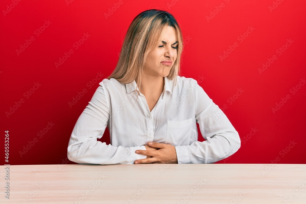 Young caucasian woman wearing casual clothes sitting on the table with hand on stomach because indigestion, painful illness feeling unwell. ache concept. Young caucasian woman wearing casual clothes sitting on the table with hand on stomach because indigestion, painful illness feeling unwell. ache concept.