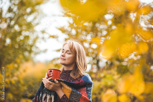 A beautiful blonde woman drinks a hot drink from a red mug in an autumn park. The woman is wrapped in a warm plaid scarf.