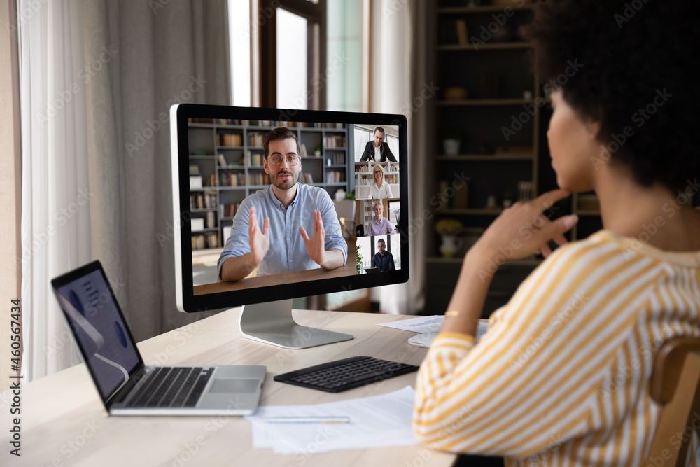 Focused young african woman looking at computer monitor, listening to skilled male team leader at brainstorming meeting with diverse colleagues using video conference call application at home office.