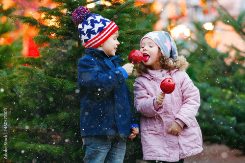 Two little smiling kids, preschool boy and girl eat sweet sugared apple on German Christmas market. Happy siblings children with lights on background and xmas trees. Family funny brother and sister.