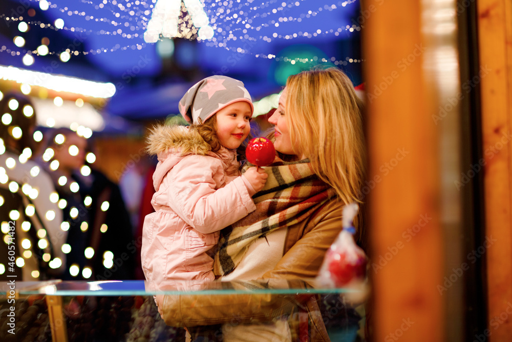 Cute little preschool girl and young mother eating crystallized sugared apple on German Christmas market. Happy family, girl and woman with lights on background. Mum holding daughter on arms. Cute little preschool girl and young mother eating crystallized sugared apple on German Christmas market. Happy family, girl and woman with lights on background. Mum holding daughter on arms.