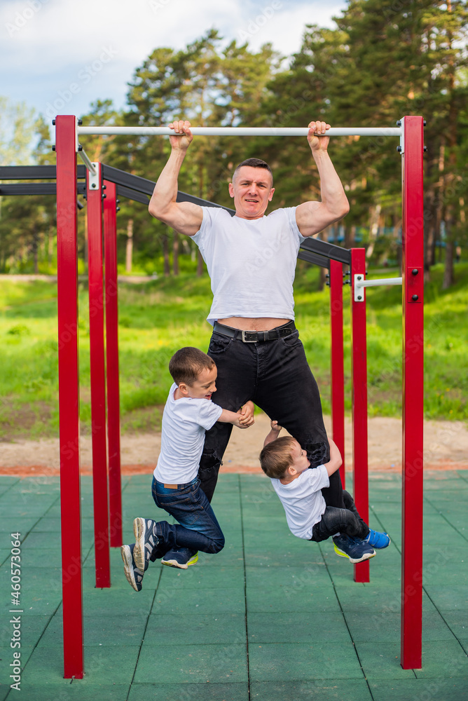 Caucasian man and two boys doing exercises outdoors. The father pulls himself up on the horizontal bar with his sons on the playground. Caucasian man and two boys doing exercises outdoors. The father pulls himself up on the horizontal bar with his sons on the playground.