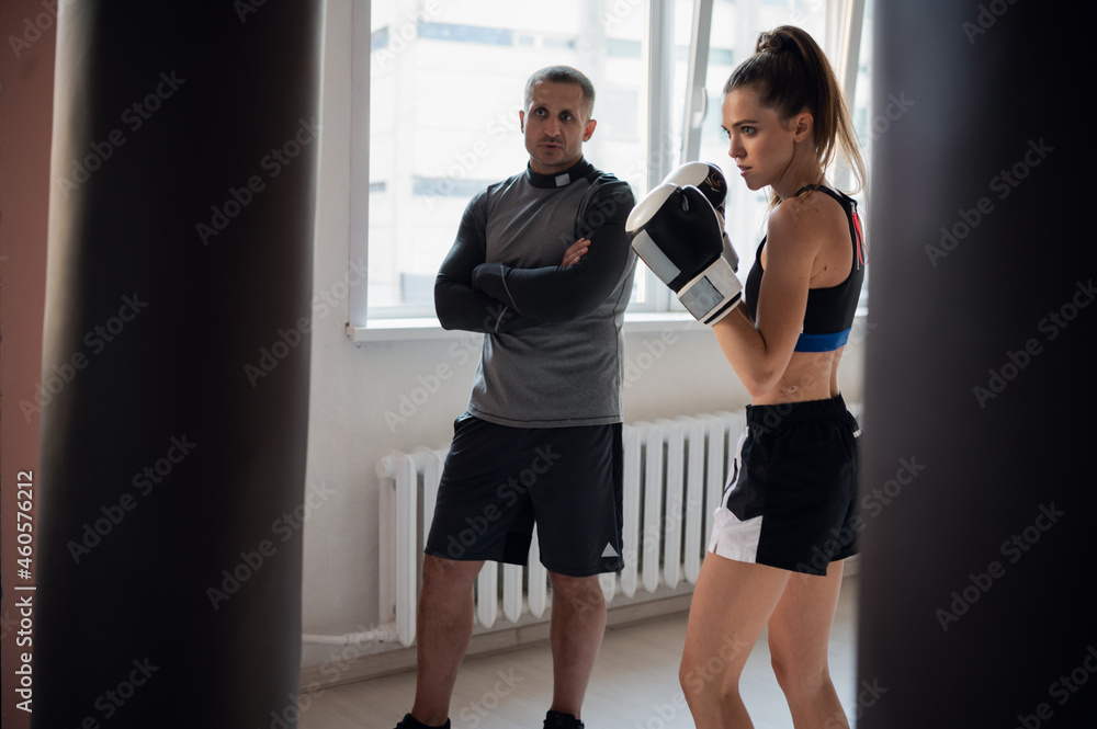 Fototapeta premium A strict trainer watches his female kickboxer student practice a punch on a punching bag in a spacious training room