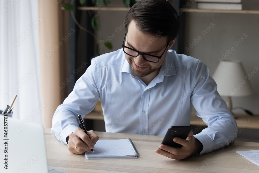 Smiling businessman in glasses taking notes in notebook, holding smartphone, sitting at home office desk, confident successful man entrepreneur writing information in personal daily planner Smiling businessman in glasses taking notes in notebook, holding smartphone, sitting at home office desk, confident successful man entrepreneur writing information in personal daily planner