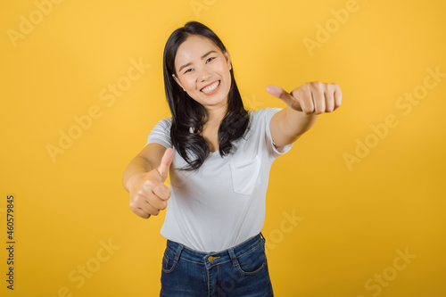 Portrait of happy, cheerful Asian woman wears white t-shirt keeps thumbs up on yellow background