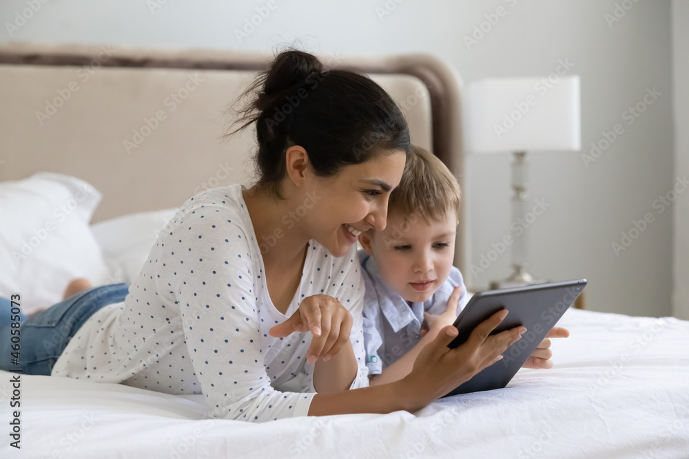 Caring smiling young indian woman using digital computer tablet applications with happy small preschool kid son, playing games resting on bed, parental control, tech addiction concept. Caring smiling young indian woman using digital computer tablet applications with happy small preschool kid son, playing games resting on bed, parental control, tech addiction concept.
