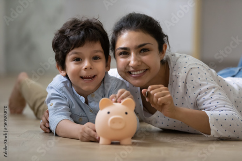 Portrait of happy indian ethnic family lying on floor with small piggybank. Caring young asian mum teaching little child son saving money for future or planning purchases, financial education concept.