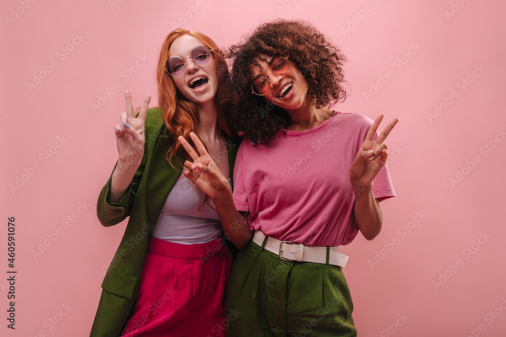 Happy cheerful young girl friends, brunette and redhead show peace sign in empty room. Smiling international women in fuchsia and emerald robes are having fun. Mood, lifestyle, concept Happy cheerful young girl friends, brunette and redhead show peace sign in empty room. Smiling international women in fuchsia and emerald robes are having fun. Mood, lifestyle, concept