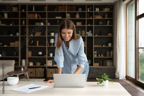 Happy millennial employee girl using laptop, standing at desk with computer, smiling at screen, typing, reading email, chatting online, making video call. Young business woman working frim home office