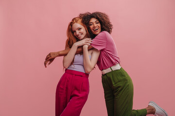Brunette with afro hair hugs embarrassed girlfriend with red hair on pink background. Beauties in casual bright clothes express positive emotions. Beauty, people emotions and summer vacation concept.