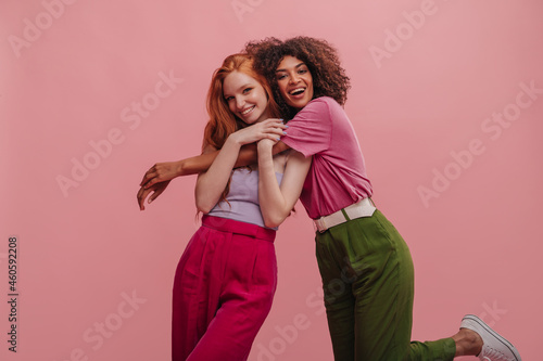 Fotografia Brunette with afro hair hugs embarrassed girlfriend with red hair on pink background
