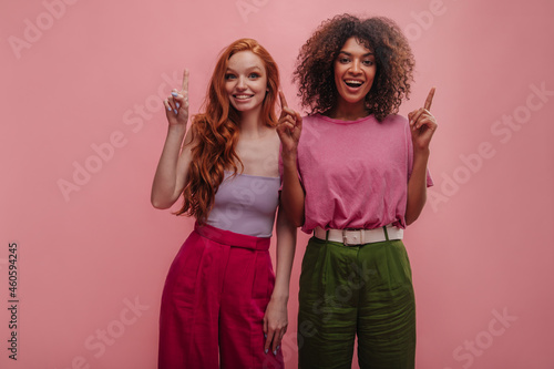 Funny young interracial girls point index fingers up with copy space. European redhead lady in tight top and trousers poses with african brunette with voluminous hairstyle in T-shirt, loose pants.