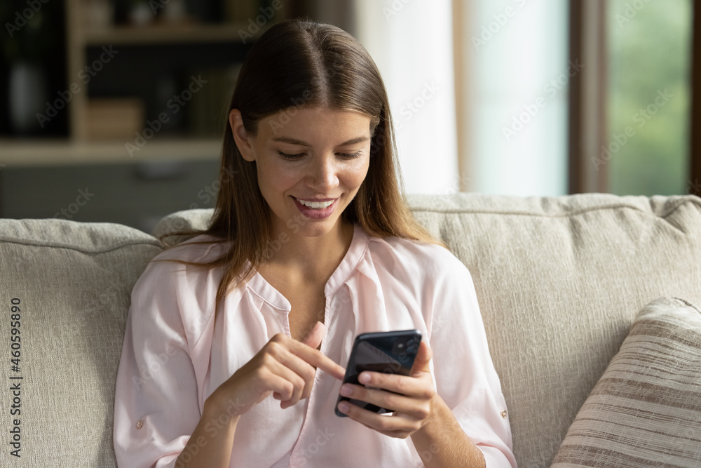 Smiling focused young woman chatting on internet on mobile phone, reading text message, making virtual order, shopping on web stores, using app for online payment, sitting on couch at home Smiling focused young woman chatting on internet on mobile phone, reading text message, making virtual order, shopping on web stores, using app for online payment, sitting on couch at home