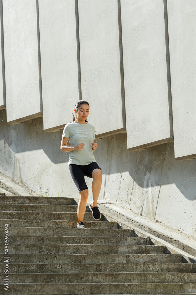 Asian woman in tracksuit runs down underground crossing large stone stairs past wall with panels training on city street in summer morning