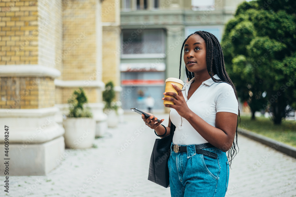 Fototapeta premium Beautiful african american woman using a smartphone and drinking coffee takeaway