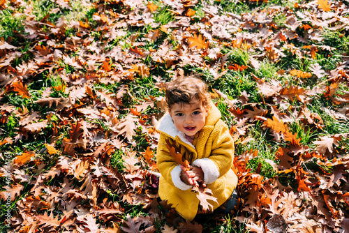 Happy little girl in yellow jacket playing with autumn leaves in park. Child in outdoor fun in fall season. Smiling cute toddler trowing fallen leaves in nature.