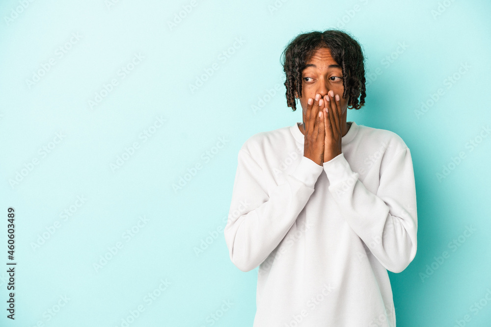 Young african american man isolated on blue background thoughtful looking to a copy space covering mouth with hand. Young african american man isolated on blue background thoughtful looking to a copy space covering mouth with hand.