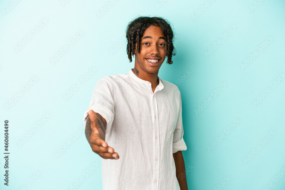 Young african american man isolated on blue background stretching hand at camera in greeting gesture. Young african american man isolated on blue background stretching hand at camera in greeting gesture.