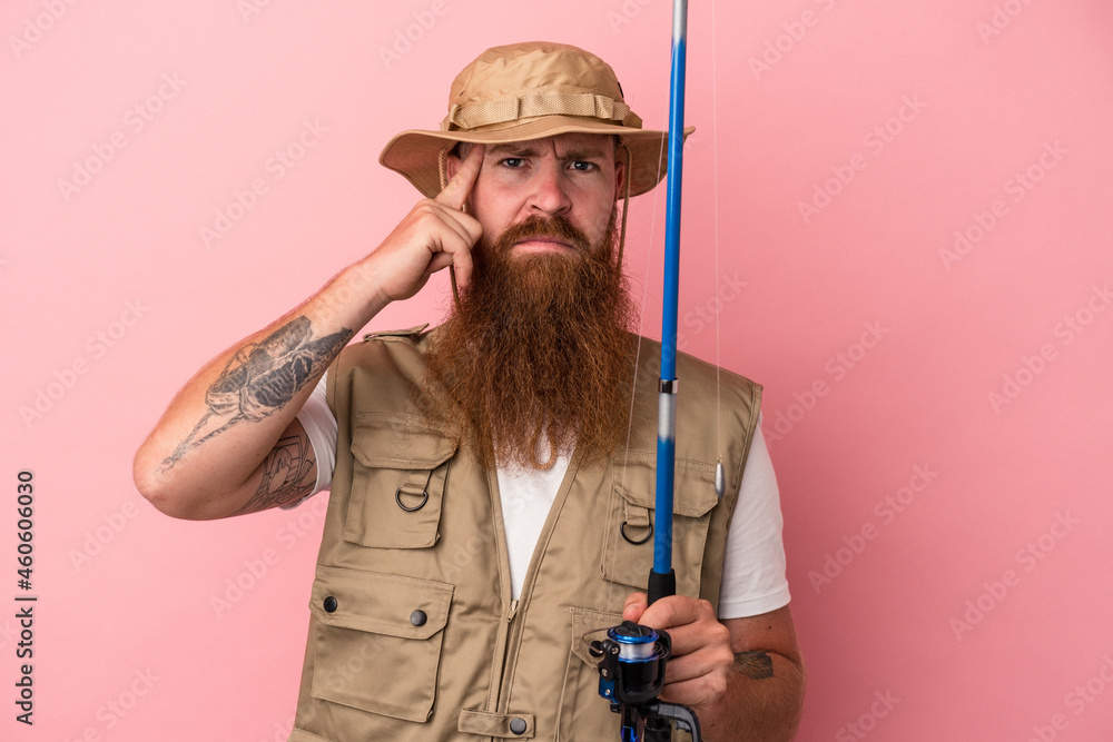 Young caucasian ginger fisherman with long beard holding a rod isolated on pink background pointing temple with finger, thinking, focused on a task. Young caucasian ginger fisherman with long beard holding a rod isolated on pink background pointing temple with finger, thinking, focused on a task.