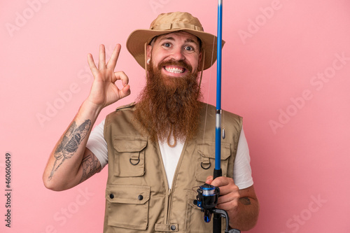 Tela Young caucasian ginger fisherman with long beard holding a rod isolated on pink background cheerful and confident showing ok gesture