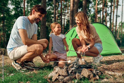 A happy family with their daughter is sitting by the campfire near the tent and frying marshmallows during the weekend in a pine forest. Camping, recreation, hiking.