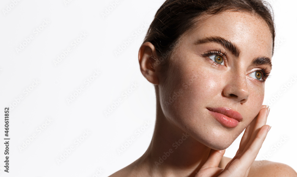 Beauty woman with freckles, thick eyebrows, touching face, using skin care lotion, anti-aging mask with detoxifying nourishing effect. Girl with naked shoulders shows clean and fresh facial skin
