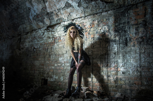 A woman in the image of a witch in a bright costume with feathers on Halloween night on the background of an old brick wall of the dungeon.