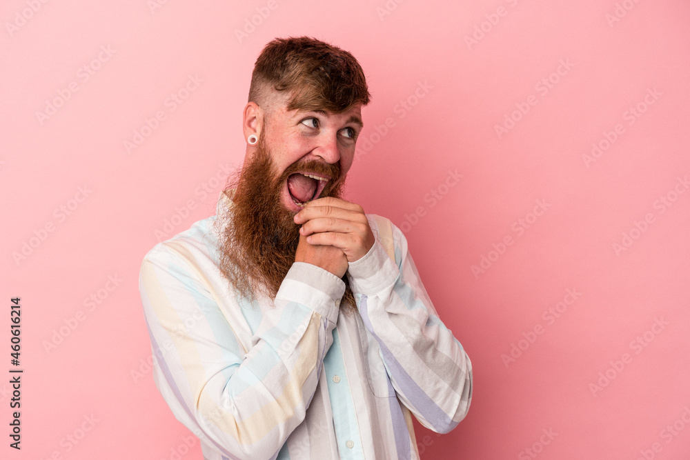 Young caucasian ginger man with long beard isolated on pink background keeps hands under chin, is looking happily aside. Young caucasian ginger man with long beard isolated on pink background keeps hands under chin, is looking happily aside.