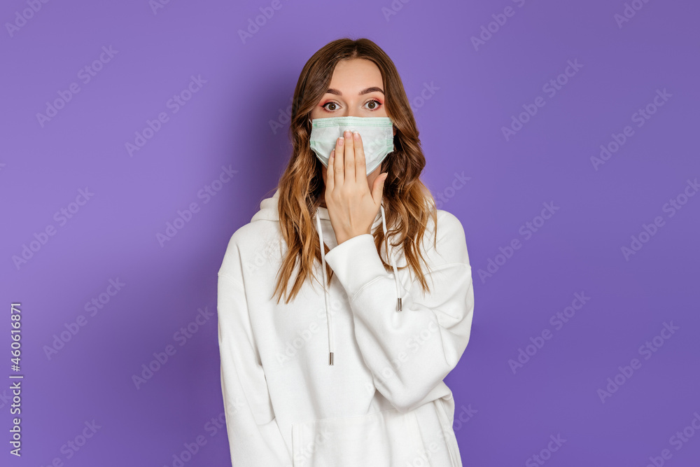 Shocked caucasian young woman wearing a medical protective mask covering her mouth with hand isolated on lilac background in studio Shocked caucasian young woman wearing a medical protective mask covering her mouth with hand isolated on lilac background in studio