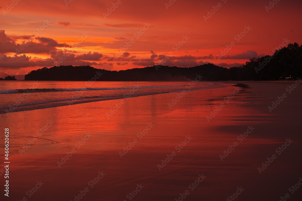 Roter Sonnenuntergang an langem Sandstrand mit flachen Wellen und Reflexionen Sillhouette