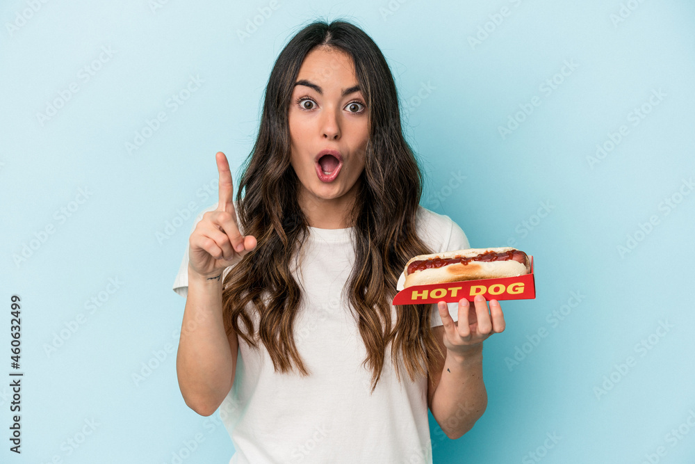 Young caucasian woman holding a hot dog isolated on blue background having an idea, inspiration concept. Young caucasian woman holding a hot dog isolated on blue background having an idea, inspiration concept.