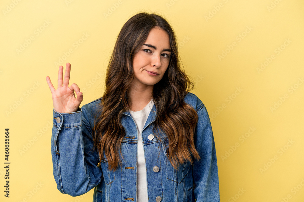Young caucasian woman isolated on yellow background winks an eye and holds an okay gesture with hand. Young caucasian woman isolated on yellow background winks an eye and holds an okay gesture with hand.