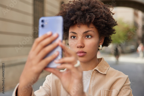 Serious Afro American woman takes photo of herself with smartphone for sharing in social networks looks attentively at camera enjoys recreation time in city. Beautiful female makes video call