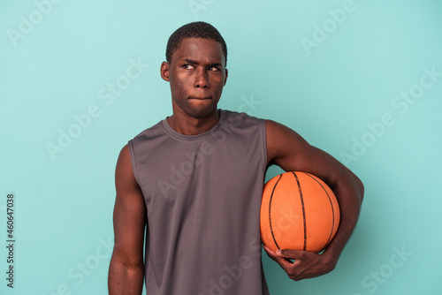 Young African American man playing basketball isolated on blue background confused, feels doubtful and unsure. © Asier