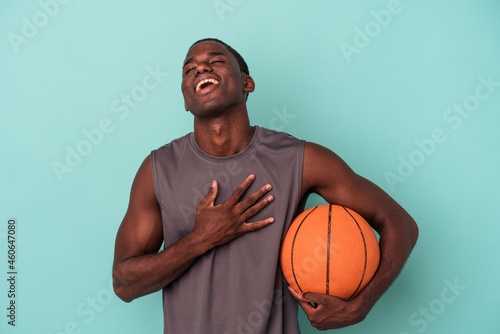 Young African American man playing basketball isolated on blue background laughs out loudly keeping hand on chest. © Asier