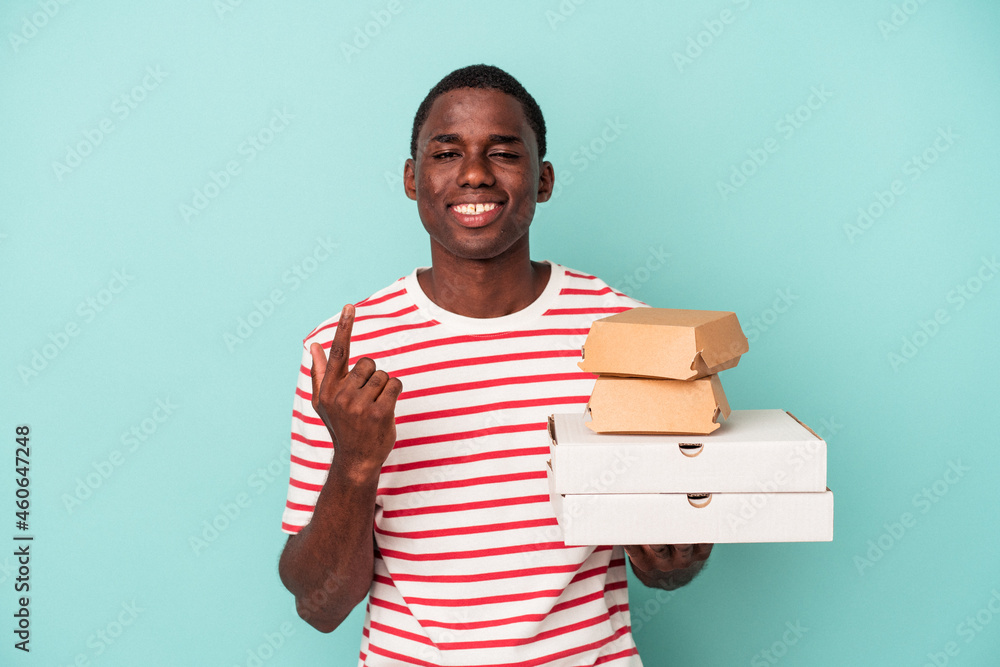 Young African American man holding pizzas and burgers isolated on blue background pointing with finger at you as if inviting come closer. Young African American man holding pizzas and burgers isolated on blue background pointing with finger at you as if inviting come closer.
