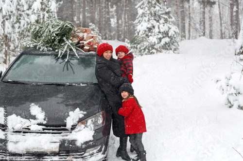 Fototapeta Naklejka Na Ścianę i Meble -  A happy mother with her daughter next to a Christmas tree from the market, loaded onto the roof of a black car. Preparation for the celebration of New Year and Christmas. Family Winter holidays