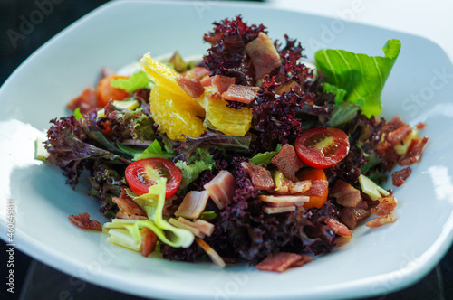 Salad with slices of egg, small cherry tomato, grilled bacon, cheese, green and purple lettuce in white bowl plate
