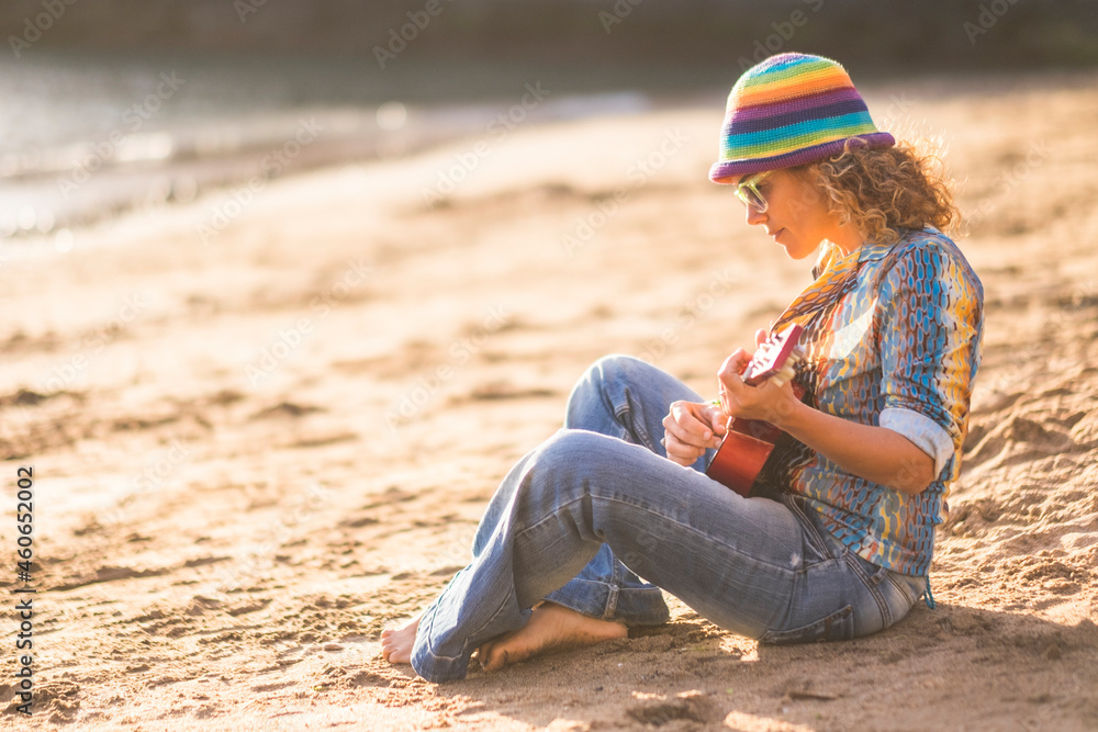 Beautiful woman sitting on sand at beach and playing guitar. Profile of stylish woman in hat and sunglasses playing guitar sitting on beach during holiday Beautiful woman sitting on sand at beach and playing guitar. Profile of stylish woman in hat and sunglasses playing guitar sitting on beach during holiday