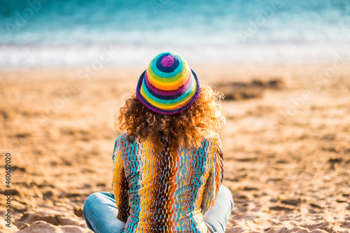 Rear view of lonely woman sitting on sand at beach. Woman relaxing peacefully while sitting on beach. Back of woman in hat admiring beautiful seascape sitting on sand © simona
