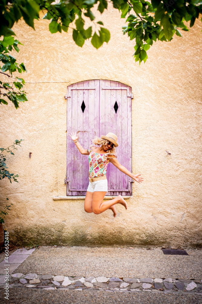 Carefree beautiful woman in hat jumping midair in front of old wooden closed window of textured building wall. Ecstatic hipster woman jumping in air outdoors during vacation