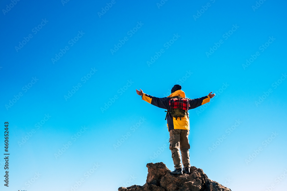 Rear view of man in backpack standing on rock with arms stretched against blue sky. Man exploring nature during vacation. Rear view of carefree man backpacker with arms outstretched against blue sky Rear view of man in backpack standing on rock with arms stretched against blue sky. Man exploring nature during vacation. Rear view of carefree man backpacker with arms outstretched against blue sky