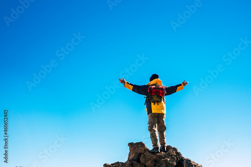 Rear view of man in backpack standing on rock with arms stretched against blue sky. Man exploring nature during vacation. Rear view of carefree man backpacker with arms outstretched against blue sky