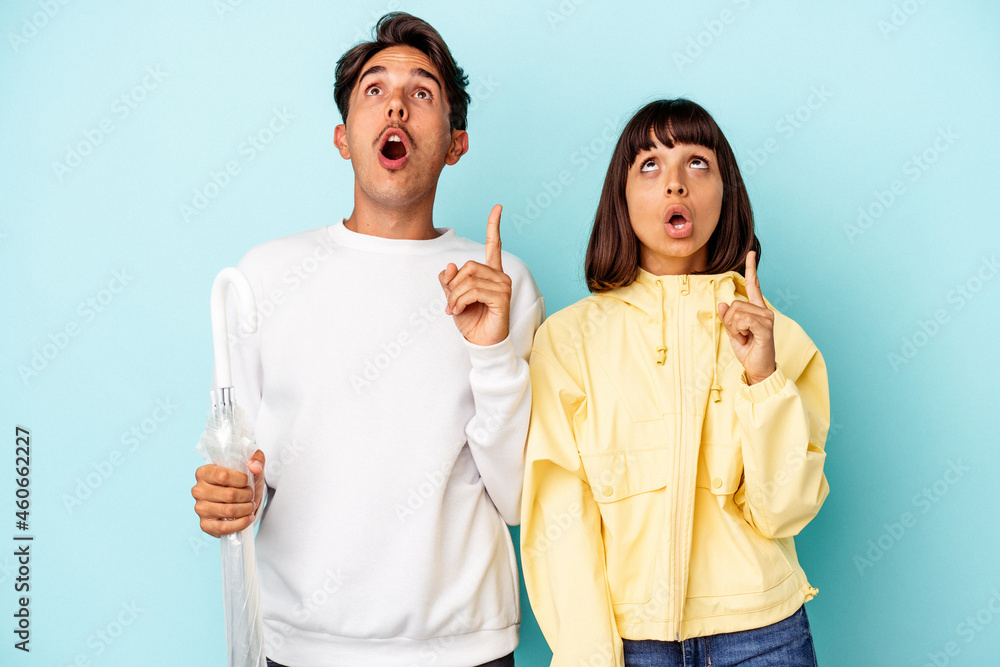 Young mixed race couple holding umbrella isolated on blue background pointing upside with opened mouth. Young mixed race couple holding umbrella isolated on blue background pointing upside with opened mouth.