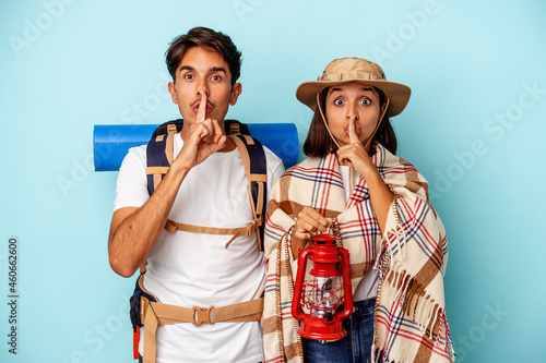 Young mixed race hiker couple isolated on blue background keeping a secret or asking for silence.