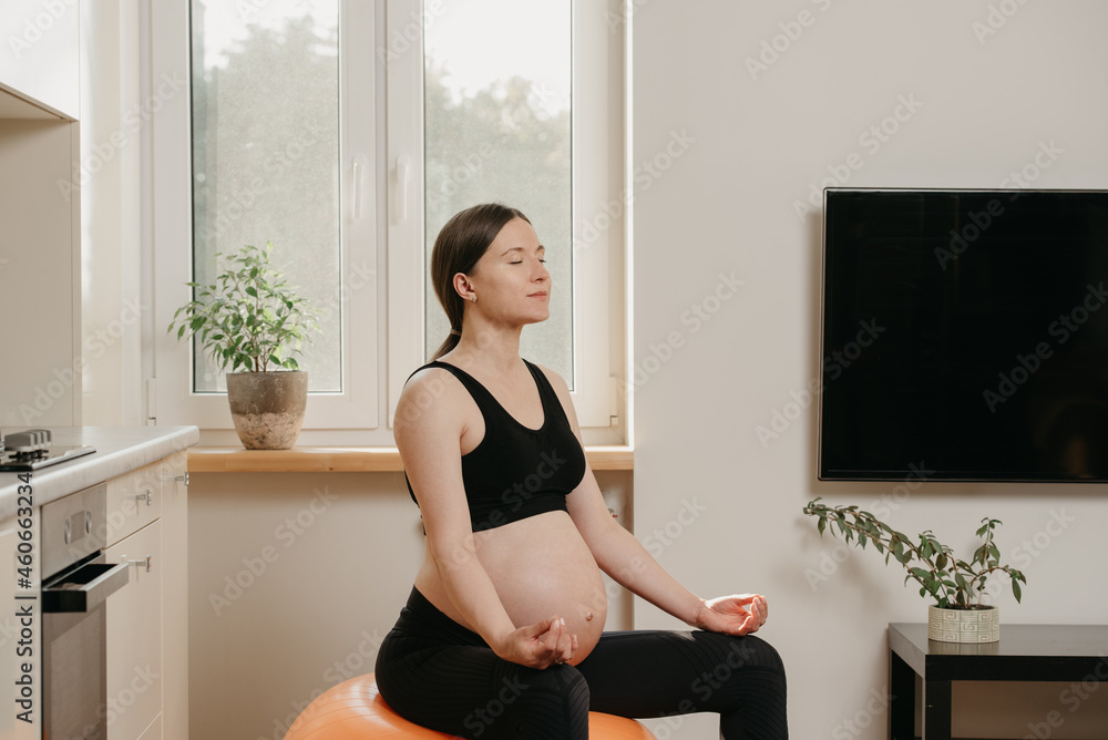 A portrait from the side of a pregnant woman who is meditating on an orange exercise ball and relaxing after a fitness training near the kitchen at her home. Healthy fit pregnancy. A portrait from the side of a pregnant woman who is meditating on an orange exercise ball and relaxing after a fitness training near the kitchen at her home. Healthy fit pregnancy.