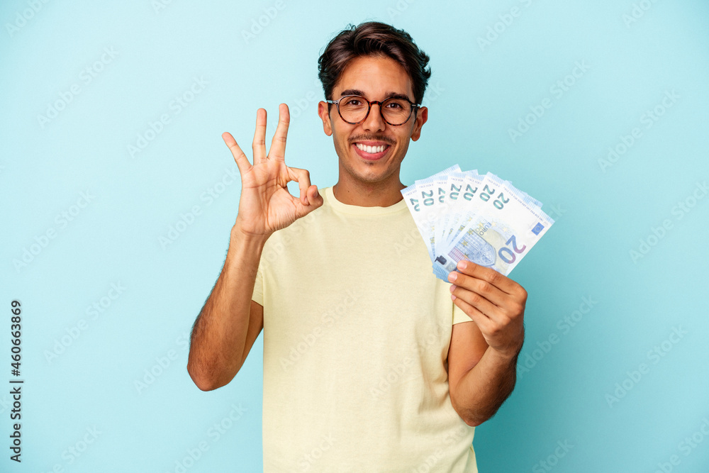 Young mixed race man holding bills isolated on blue background cheerful and confident showing ok gesture. Young mixed race man holding bills isolated on blue background cheerful and confident showing ok gesture.
