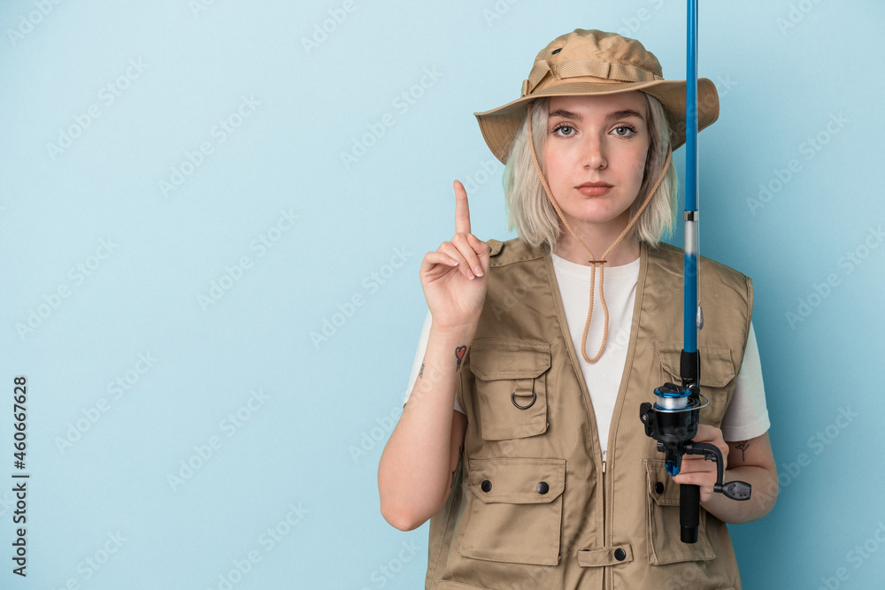 Young caucasian fisherwoman holding a rod isolated on blue background showing number one with finger. Young caucasian fisherwoman holding a rod isolated on blue background showing number one with finger.