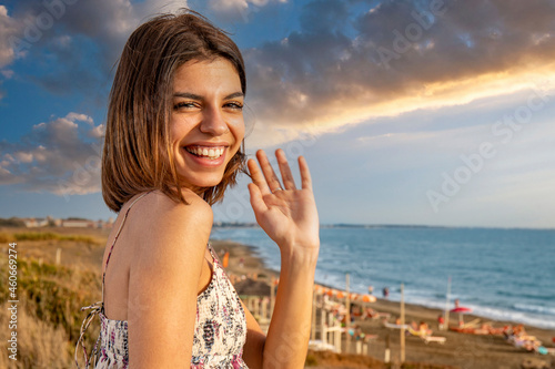 A young smiling woman waves at the spectator. Cheerful gesture and happy air. A seaside landscape at the sunset in the background.